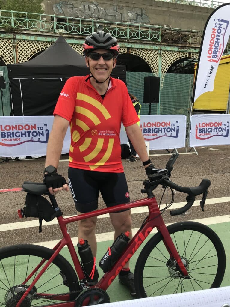 A man in a red shirt with a yellow rotor graphic on it stands with his bike at the end of the London to Brighton cycle ride.