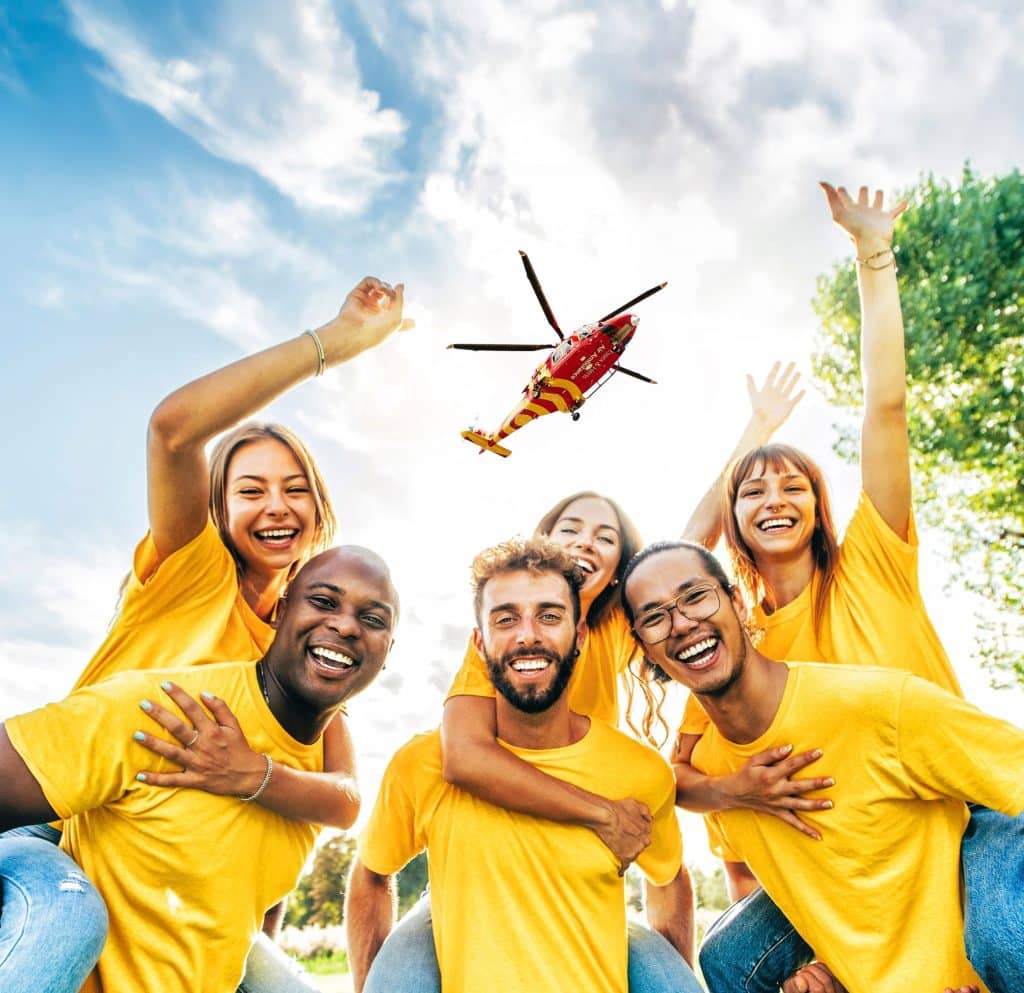 A group of racially diverse young people smiling in matching yellow shirts with an EHAAT helicopter flying in the sky above them