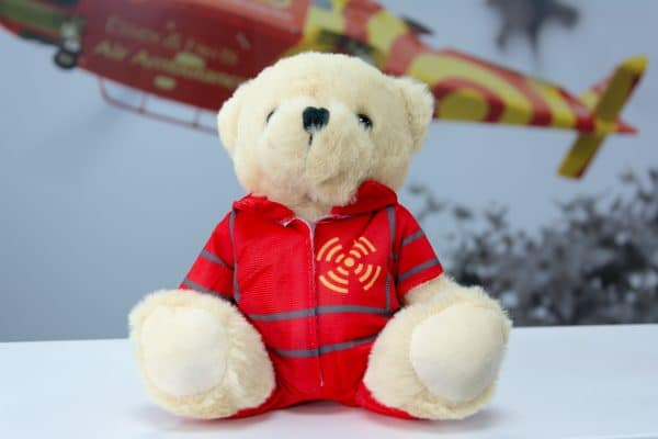 Golden teddy bear wearing an Essex & Herts Air Ambulance flight suit in front of large format photo of the AW169 in helicopter in flight